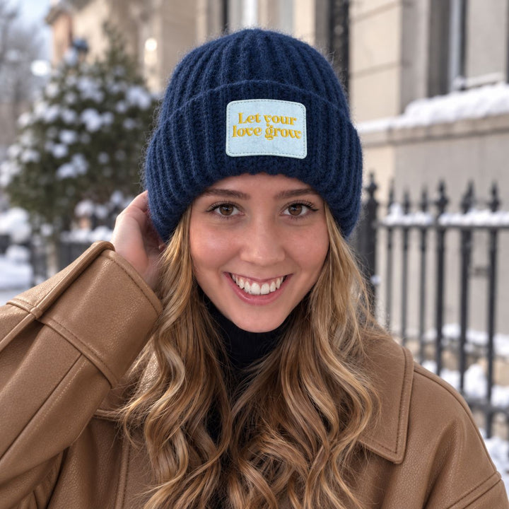 Blonde Frau trägt navyfarbene CASHIMAR Mütze mit „Let your love grow“-Patch und camel-farbene Jacke vor winterlicher Stadtkulisse.