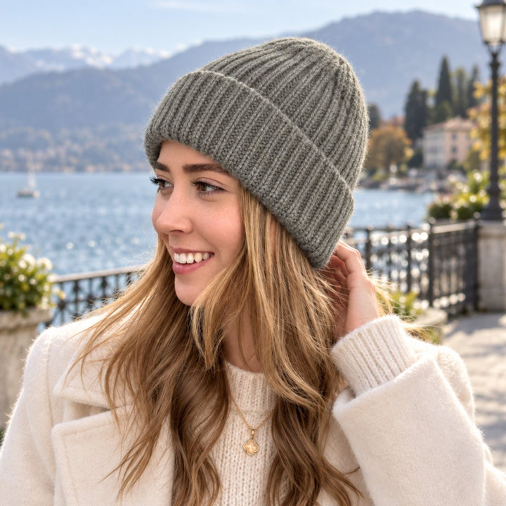 Luana trägt die 100% Cashmere Mütze Bari in Seagrass, einer graugrünlichen Farbe, kombiniert mit einem cremefarbenen Mantel in einem stilvollen Outdoor-Setting an der Seepromenade.