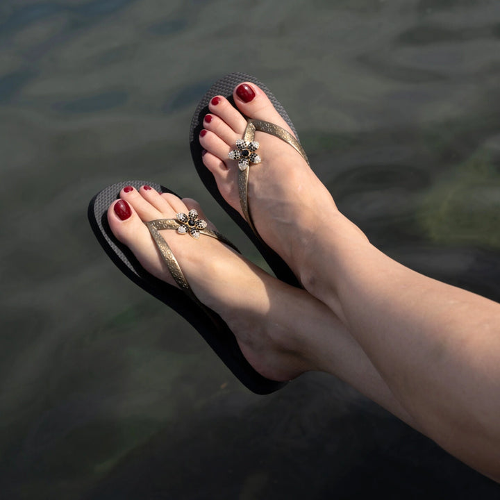 Simone Herrera Chanclas Yasmin in Schwarz mit Blume, getragen am Wasser, sommerlicher Flip Flop Lifestyle-Shot.