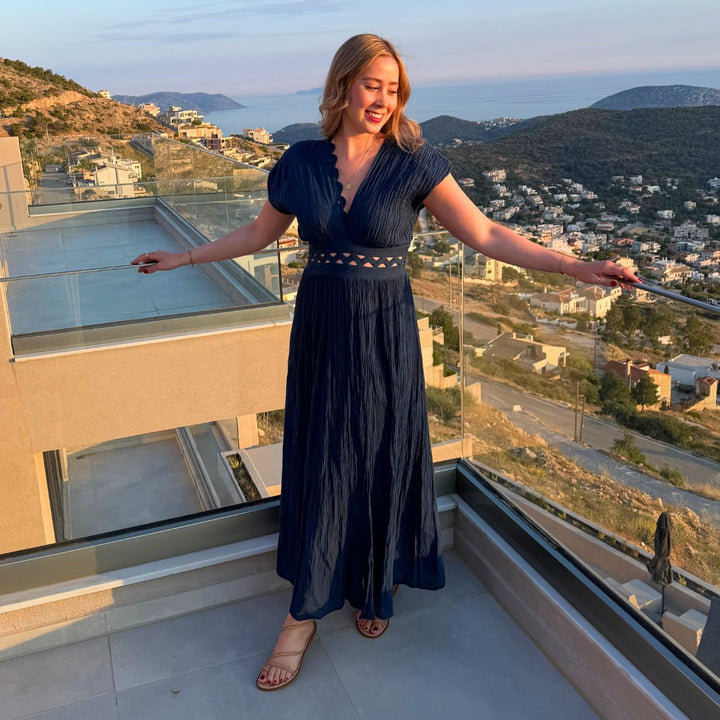 Luana im dunkelblauen D'Artemide Nerea Dress auf einer Terrasse in Griechenland mit Blick auf die Küste bei Sonnenuntergang.