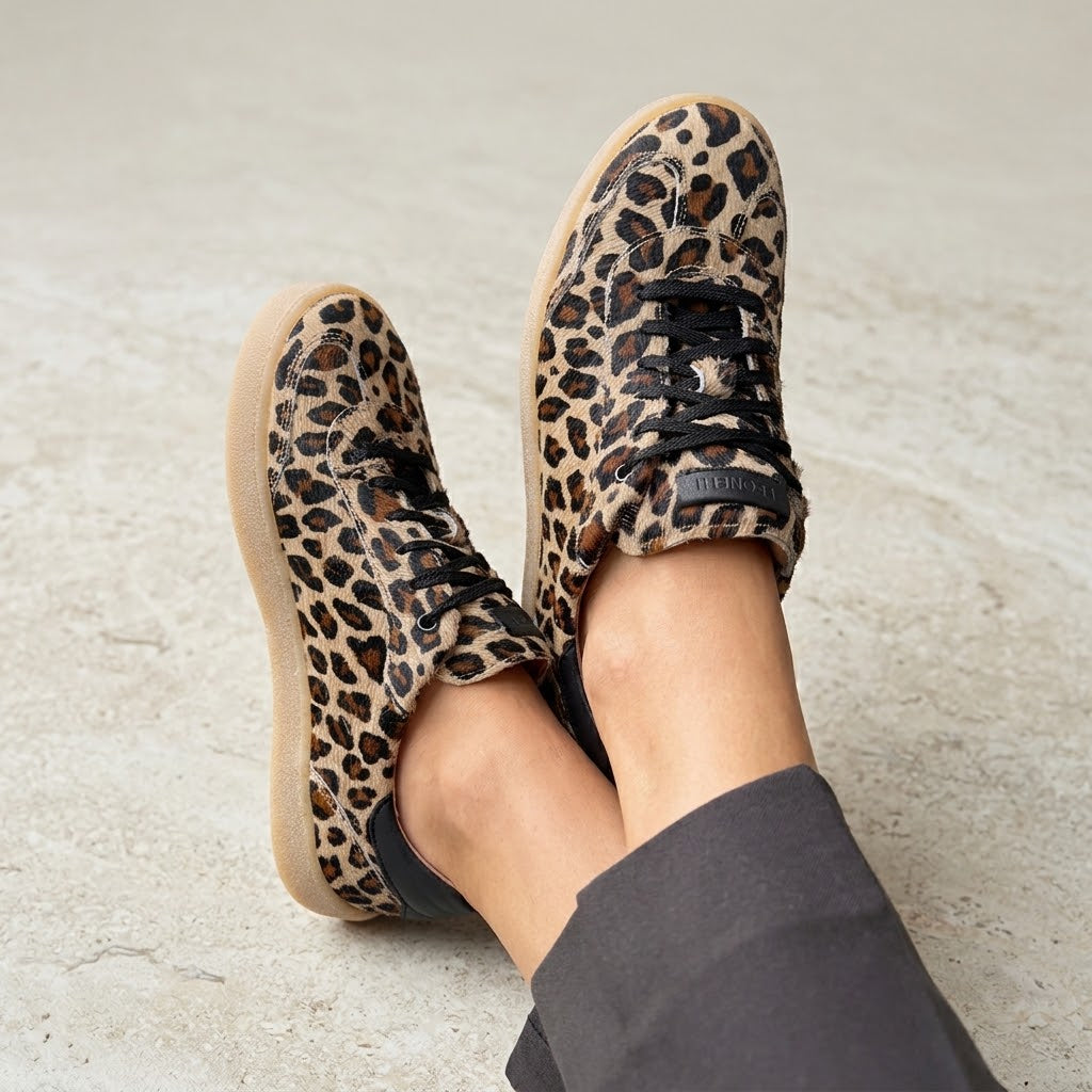 Dianetti Sneaker überkreuzt an Damenfüssen mit brauner Adele Carmine Hose auf beigem Steinboden.