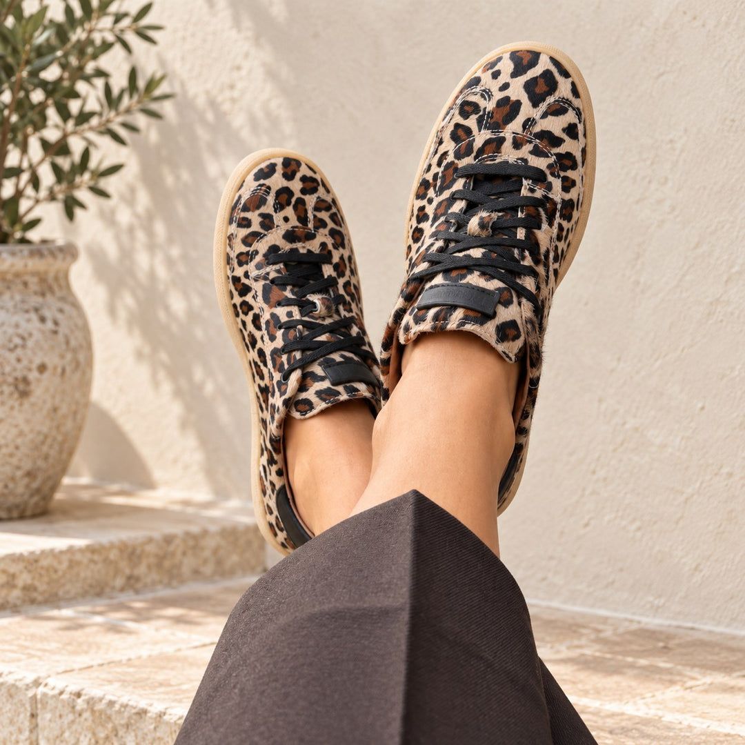 Dianetti Sneaker überkreuzt an Damenfüssen mit brauner Adele Carmine Hose im mediterranen Setting.