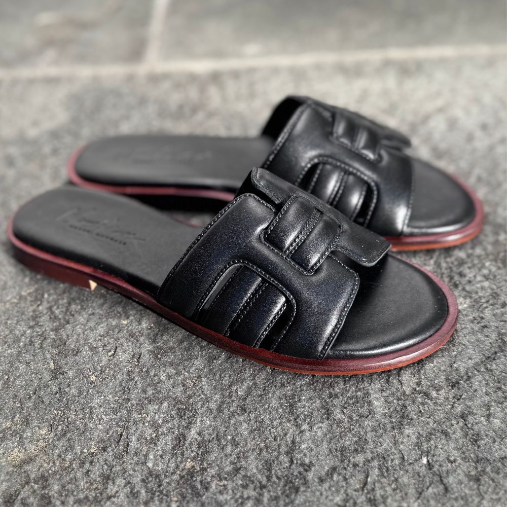 NoAna Greek Sandals City Chic black, flache Sandalen aus schwarzem Glattleder, klassisch gestylt auf Steinboden im Outdoor-Setting