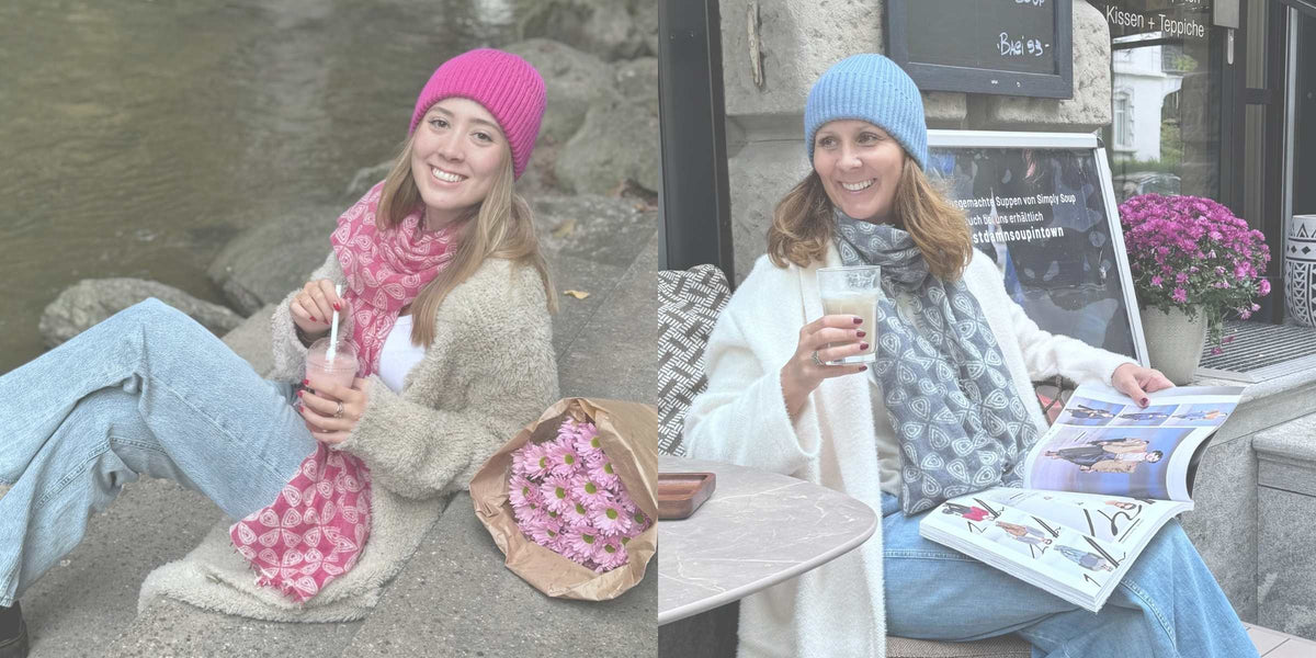 Luana in Jeans mit Schal und Mütze in pink und Janine mit Jeans und Schal und Mütze in blau mit Getränk in der Hand sitzend.